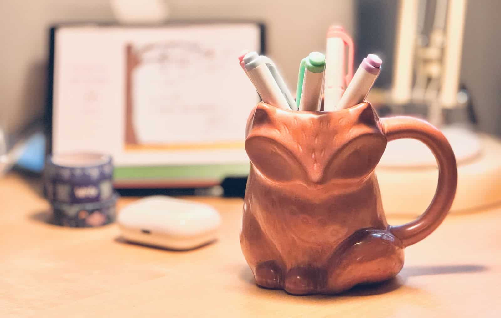 Picture of a desk showing a fox-shaped mug holding a collection of pastel highlighters, with a daily calendar, washi tape, and Airpods in the background.